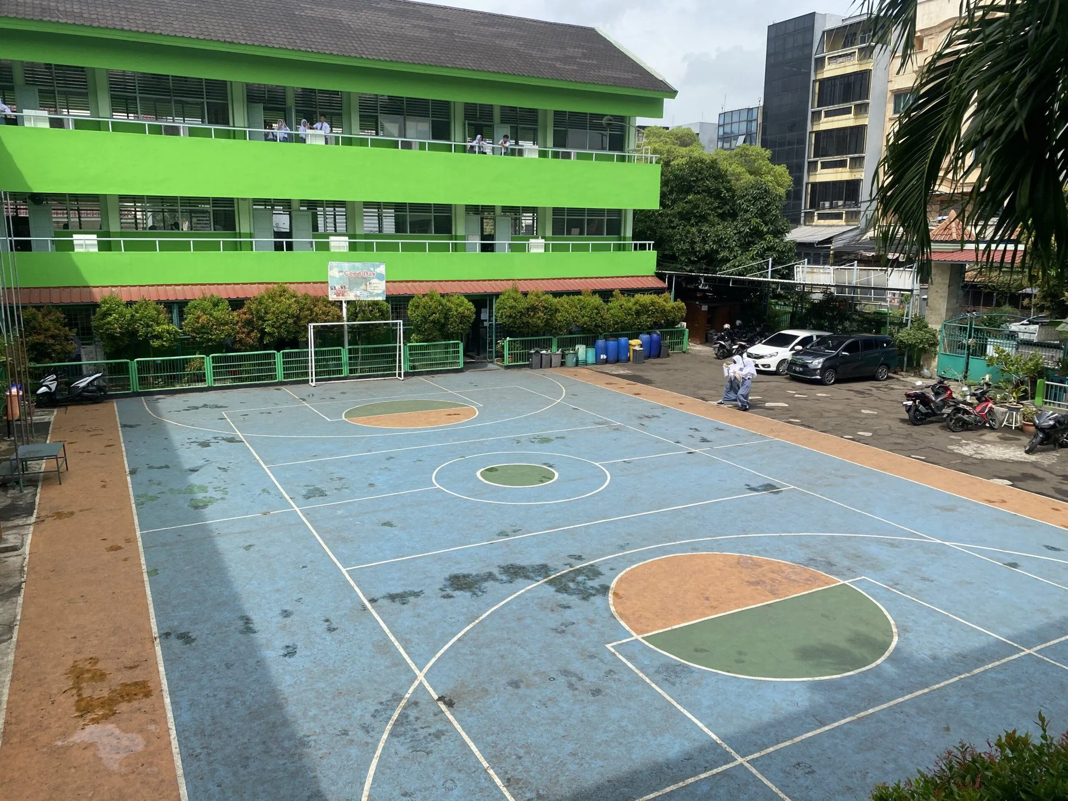 Gedung tiga lantai SMKN 11 Jakarta berwarna hijau dan putih di belakang lapangan olahraga.